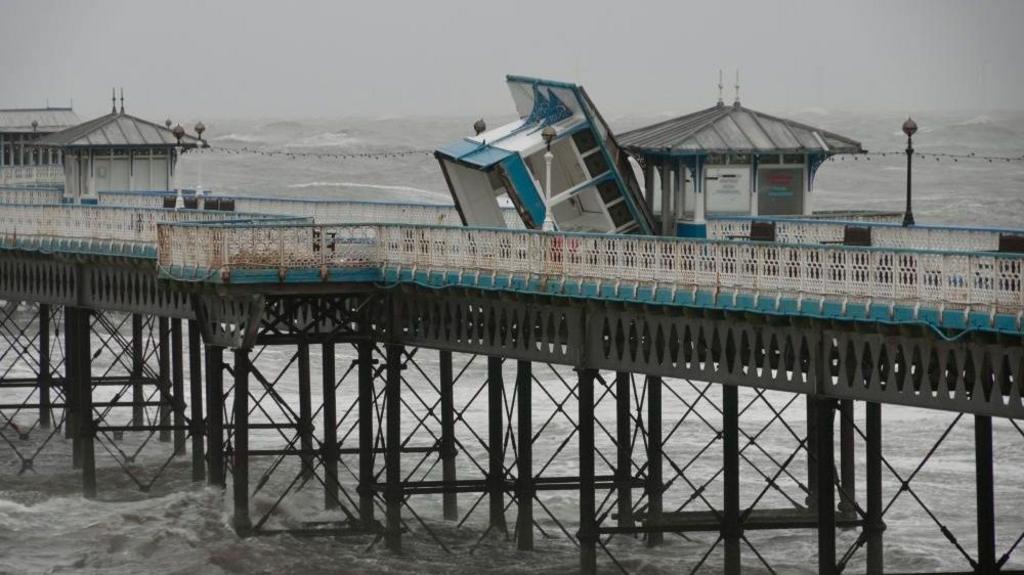 Damage to Llandudno pier, small building with benches tipped over onto the side with waves behind