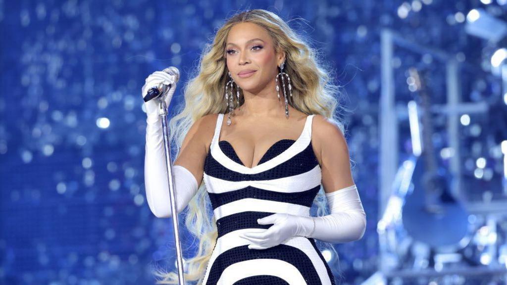 Beyonce, wearing a black and white dress with white gloves, holding a microphone on a stand on stage during her Renaissance World Tour in 2023.