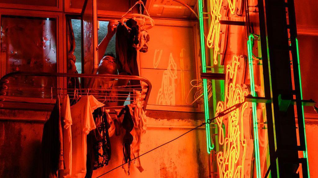 A man hangs laundry next to a lit neon sign outside the window of his flat in Hong Kong on November 18, 2024.