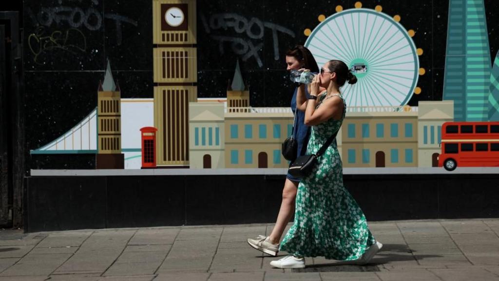 Two women walking past a painting of the London skyline, one is drinking from a large bottle of water.