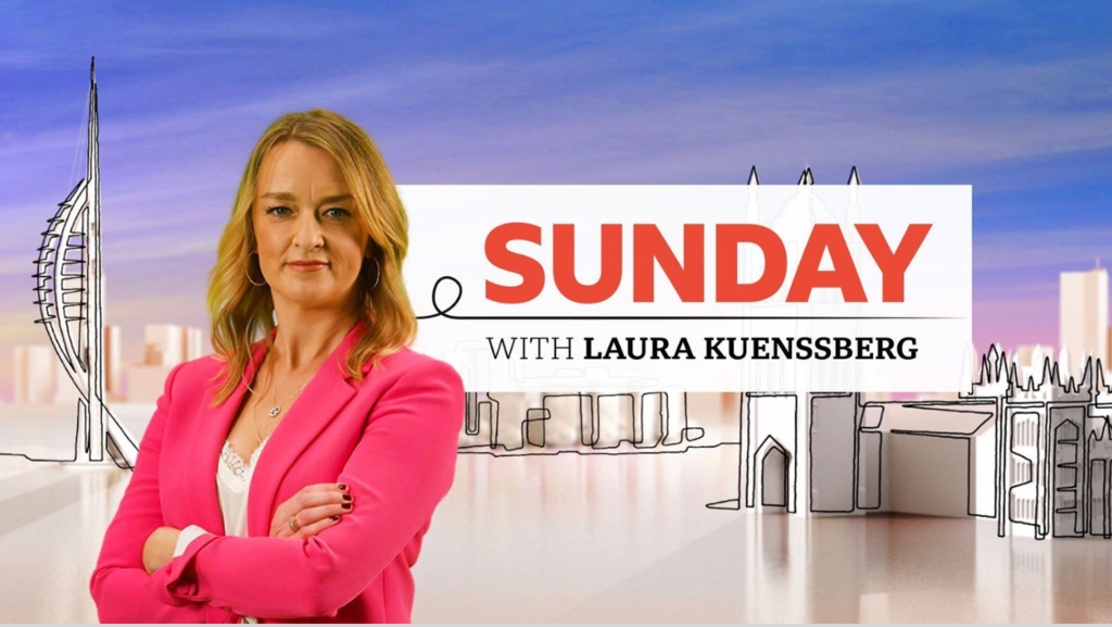 Laura Kuenssberg in pink jacket, arms folding, stood in front of branding for her show.