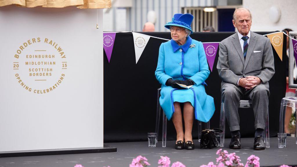 The late Queen and Prince Philip sit next to a sign marking the opening celebrations of the Borders. She is in a blue dress and coat with a blue hat and he is wearing a grey suit with a dark striped tie.