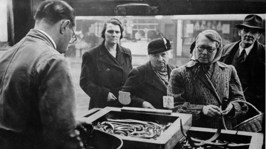 Southwark residents buying jellied eels from a stall at the Elephant and Castle in South London, 8th January 1949. Black and white photo of three women looking crossly at a box of eels