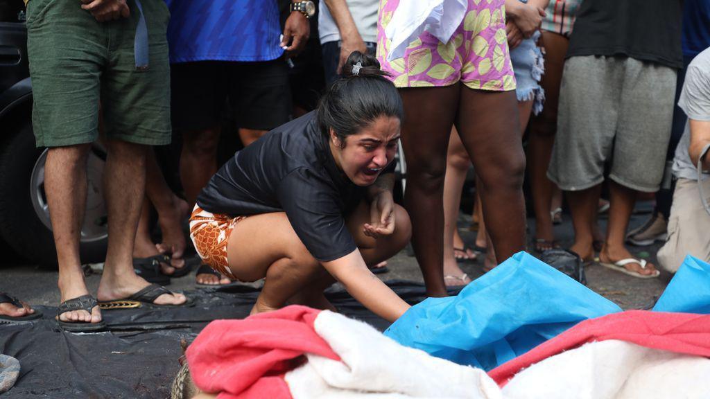 A woman lifts a blue tarpaulin which covers a body. She appears to be crying out. The legs of bystanders can be seen behind her.