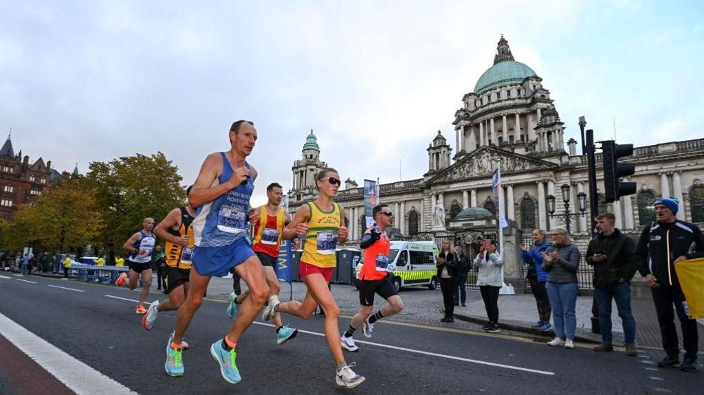 Six runners in vests and shorts, sprinting past Belfast City Hall, a large stone building with a blue, domed roof. There are a number of spectators cheering the athletes on.
