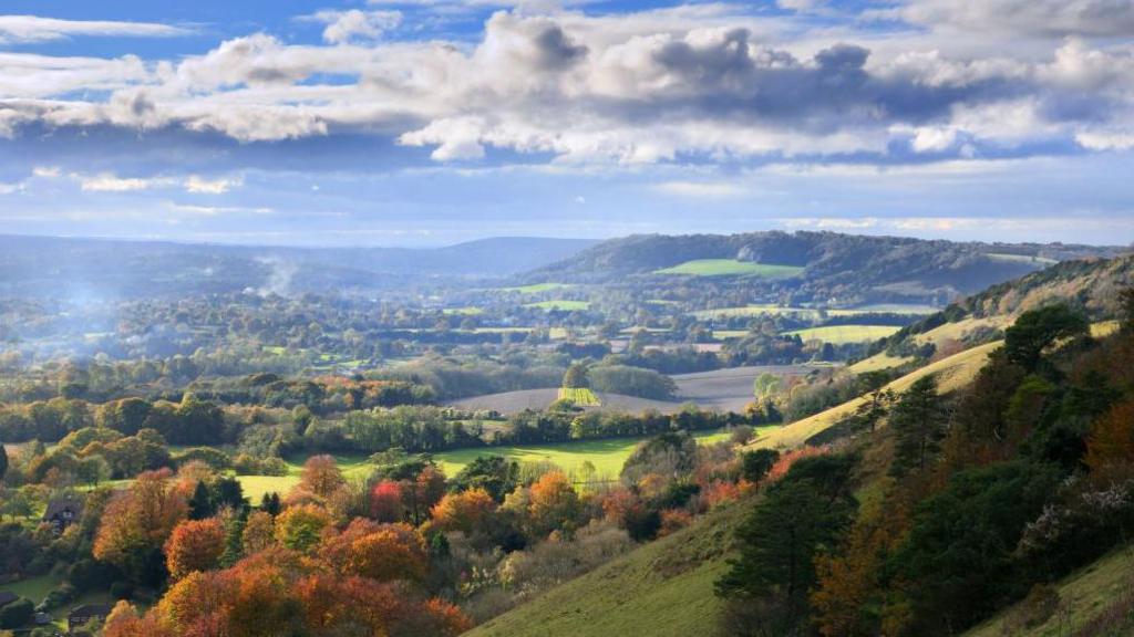 The Surrey Hills. There are lots of rolling hills with trees ranging from green, orange and red. The sky is blue and there are white clouds with the sun shining through. 