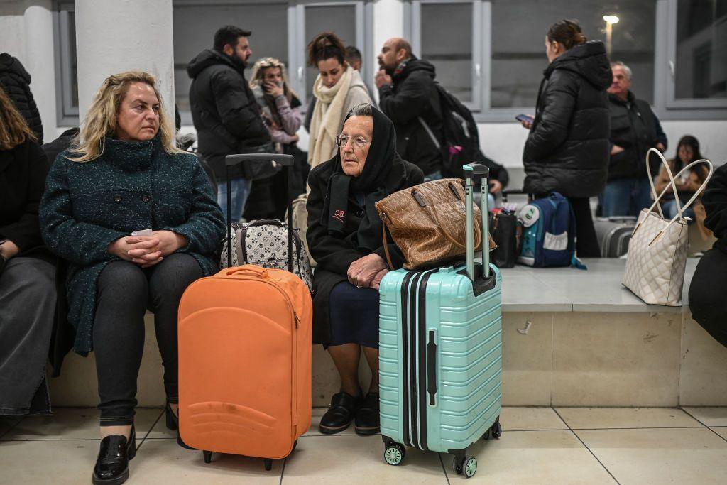 Two women sit on a bench in a ferry terminal. They both have suitcases and bags and aren't smiling. In the background there are many other people wrapped up warm and carrying bags.