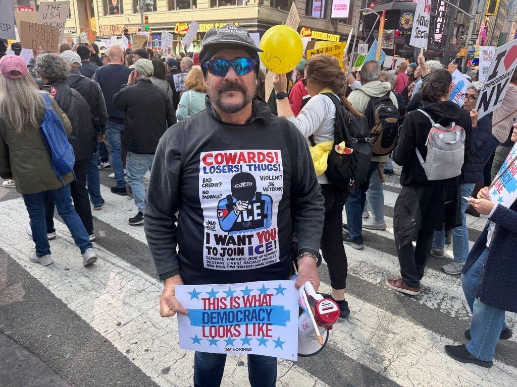 Massimo standing and smiling at camera holding sign that says "this is what democracy looks like" as crowd of protesters march behind him