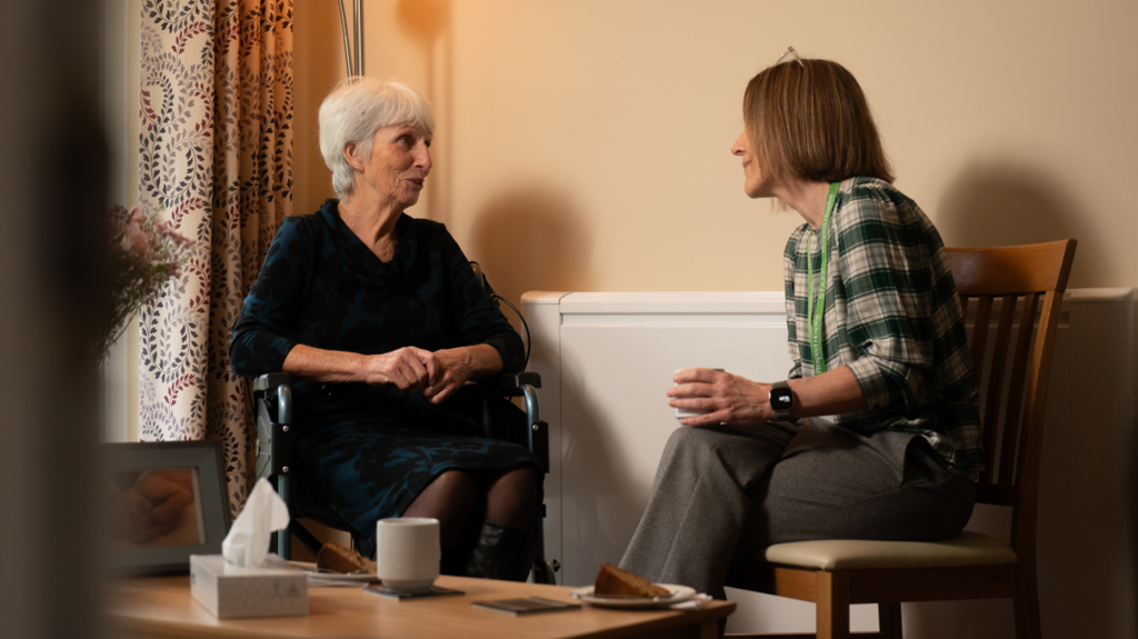 Two women chatting. They are both sitting on chairs and there is a table in front of them with a box of tissues, a cup and a plate with a slice of cake on it. One woman has short white hair, the other has brown hair in a bob haircut.