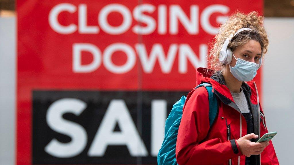 Woman wearing face mask and headphones walks past a closing down sign in a Cardiff shop window