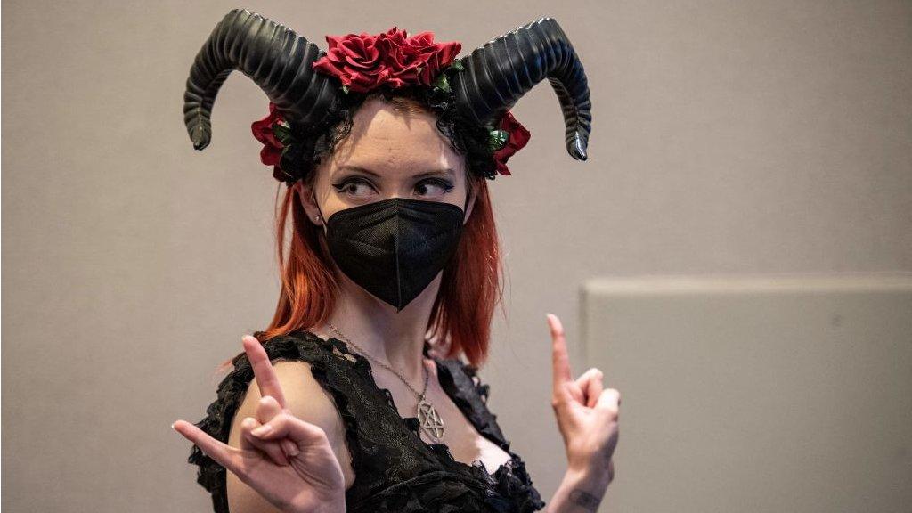 A woman wearing elaborate black horns and a Covid face mask poses for photos giving the sign of the horns at SatanCon in Boston, Massachusetts