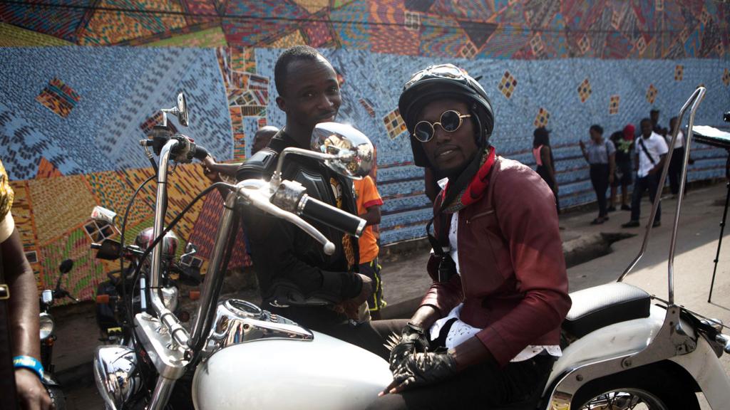 Two men sit and stand by a motorbike as they attend the Chale Wote street art festival in Accra, on 21 August 2016