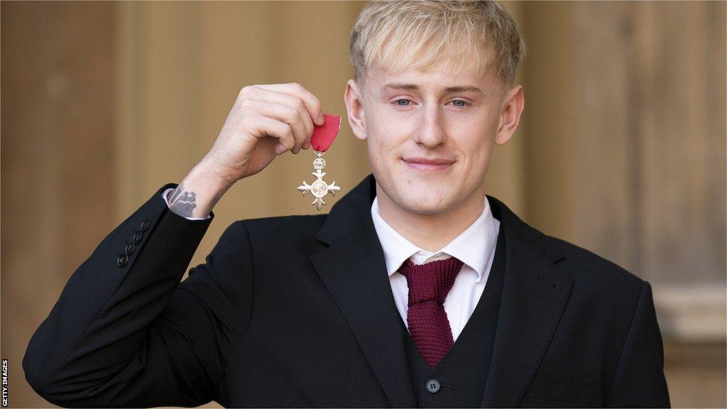 Jordan Catchpole holds up his MBE outside Buckingham Palace