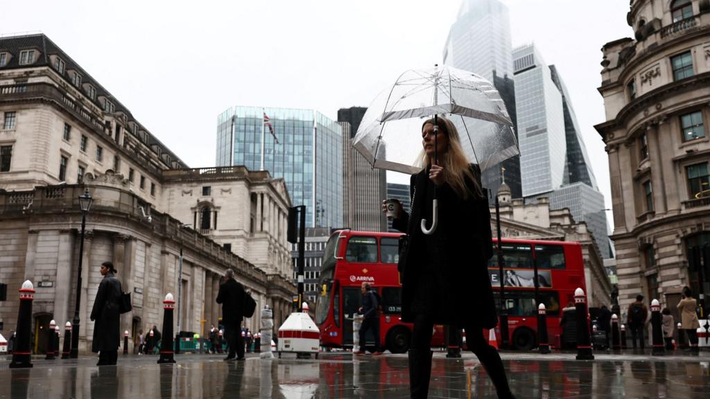 A pedestrian with an umbrella walks by the Bank of England building (L), in the financial district, central London, on November 2, 2023.