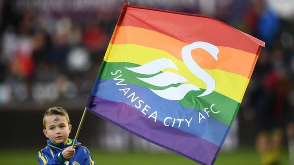 Mascot holds a flag displaying colours in support of the Stonewall Rainbow Laces campaign before the match