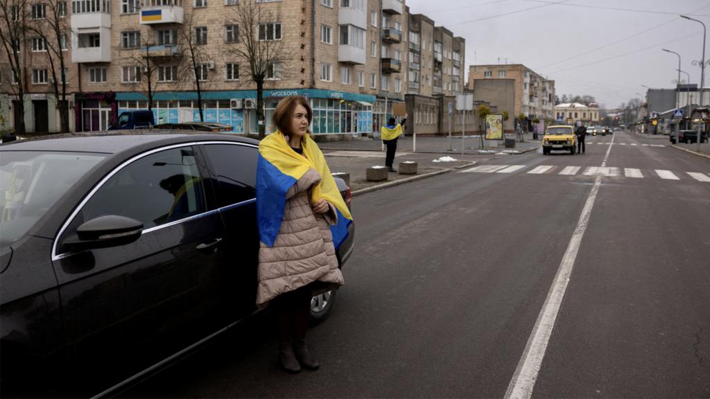 A woman draped in a Ukraine flag stands in a street next to a car as a minute silence is observed for fallen soldiers, in Kovel, Ukraine on 19 November