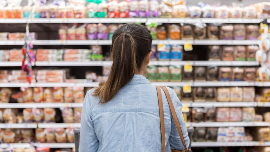 A woman pictured from behind stands with a shopping cart in front of a shelf full of food in the bread aisle of a grocery store