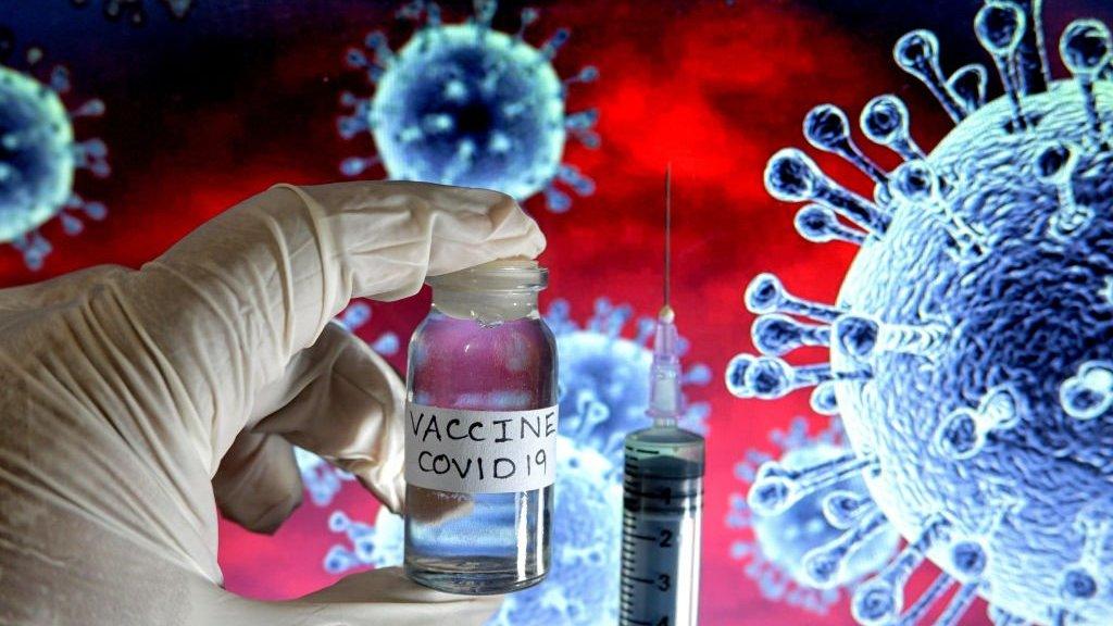 A syringe and a gloved hand holding a vial labelled "VACCINE COVID-19" with a background graphic illustration of a virus