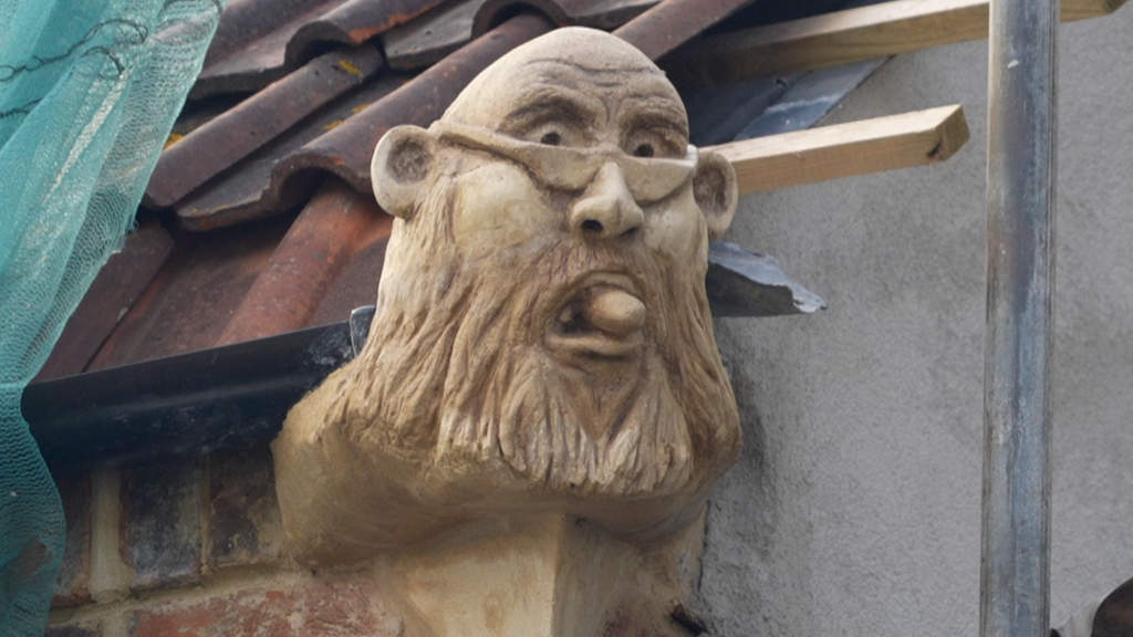 A sculpture of a man with a beard and glasses, on the edge of a roof