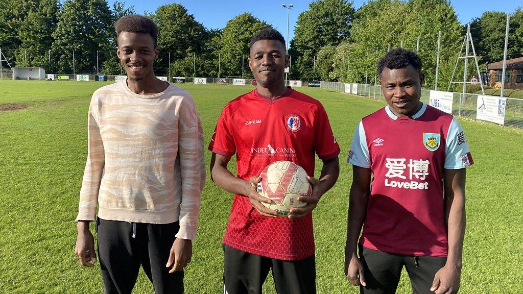 Three football players smiling stood on the edge of a football pitch on a sunny day