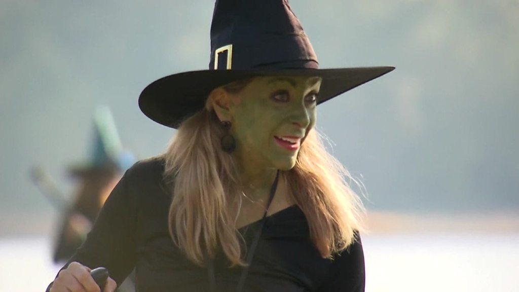 A woman wearing a witches hat and green make up on her face smiles at someone,