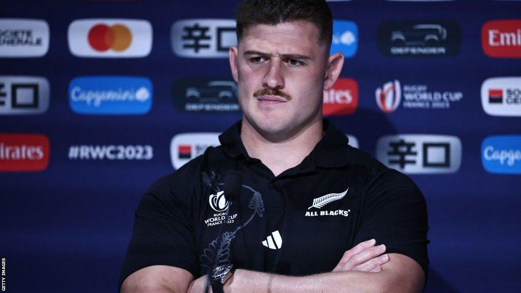 Dalton Papali'i sits arms folded behind a microphone and in front of a Rugby World Cup 2023 backdrop