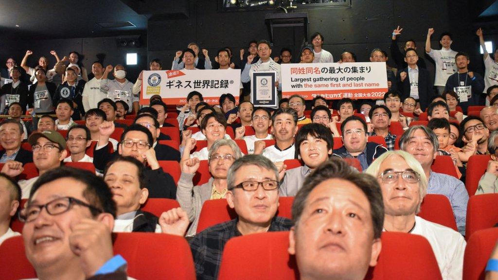 Group of men in a theatre, holding a poster reading "largest gathering of people with the same first and last name"