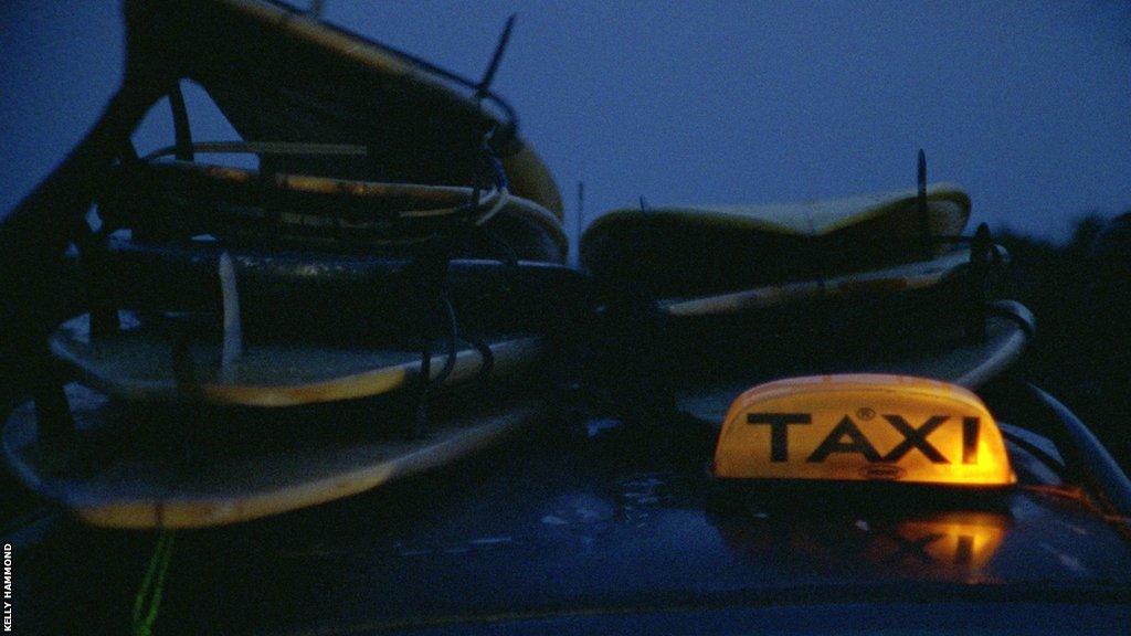 A taxi with illuminated sign is loaded with surfboards on the roof