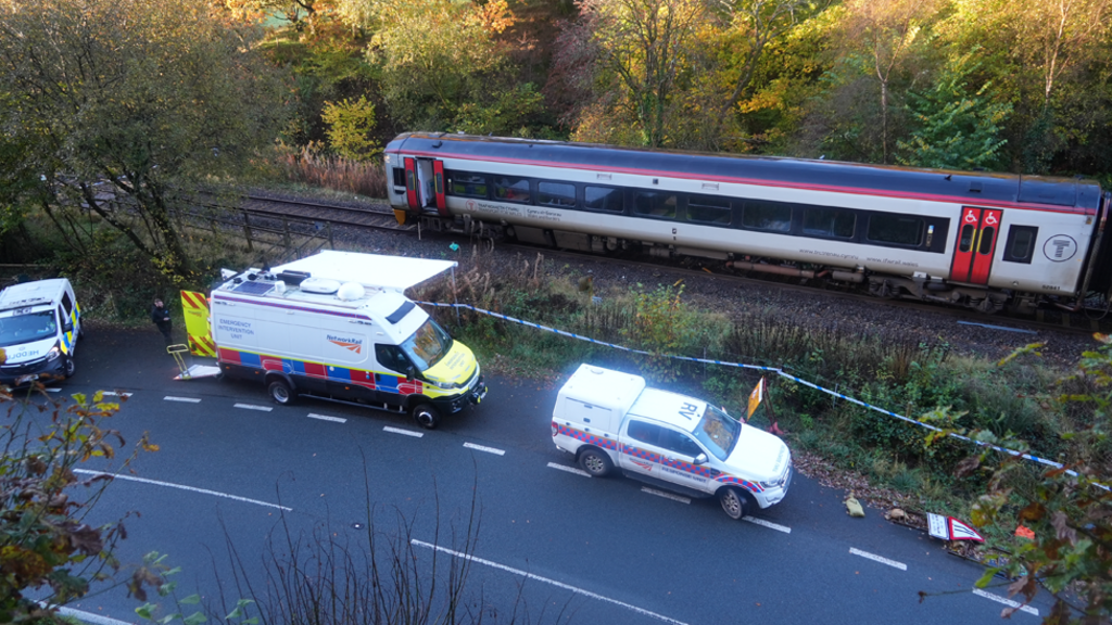 Wide shot of the train carriage with open doors stationary on the track. There are three Network Rail vehicles on the road outside and police tape in place