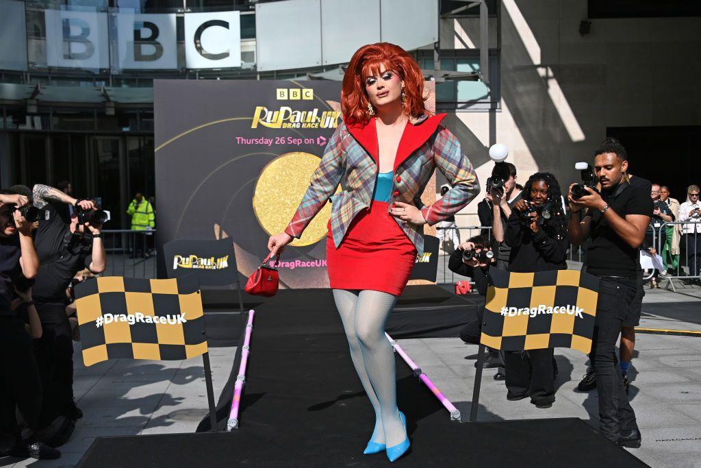 Chanel O'Connor, a drag Queen competing on this year's Ru Paul's Drag Race UK. She is wearing a red wig and lots of makeup. She has in gold earring and is wearing a tartan blazer. She is posing at the end of a catwalk at the BBC's broadcasting house in London.