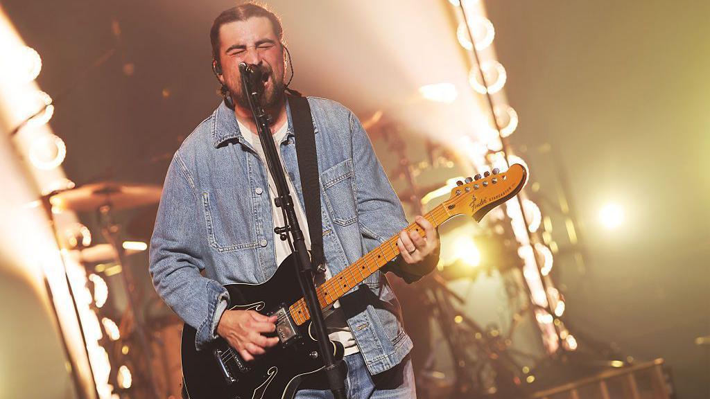 Noah Kahan singing and playing a guitar