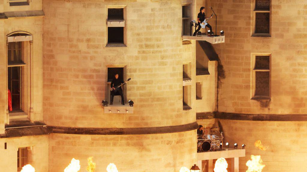 A rock band, with two guitars and drums, play from the side of a castle-like building