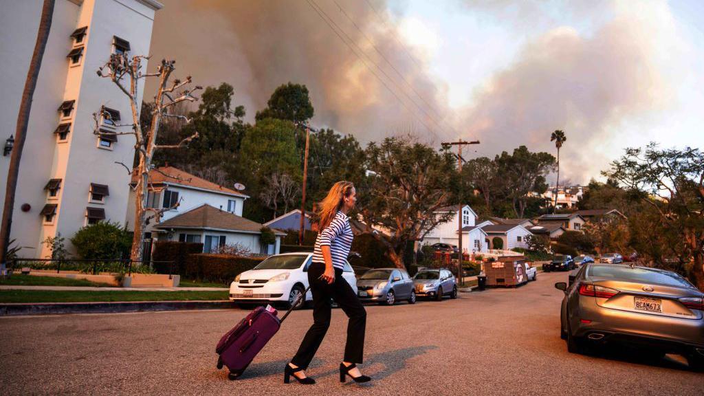 A woman rolls a suitcase to her car parked on an LA street while smoke billows in the distance