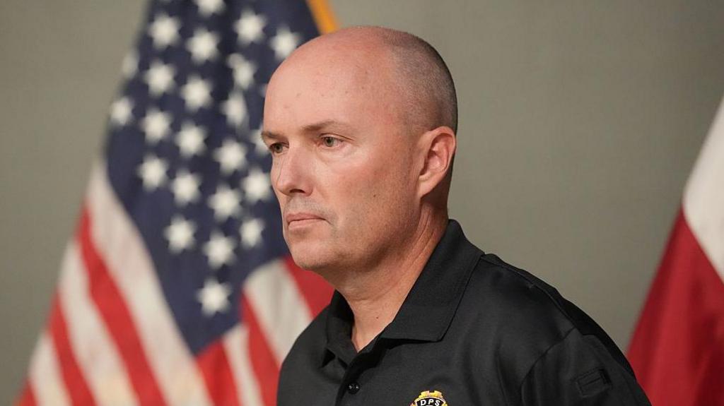 Utah Governor Spencer Cox shoulder cropped image. He is facing left and in the background is a blurred US flag on a pole to his left. To the back right is a blurred partial flag.