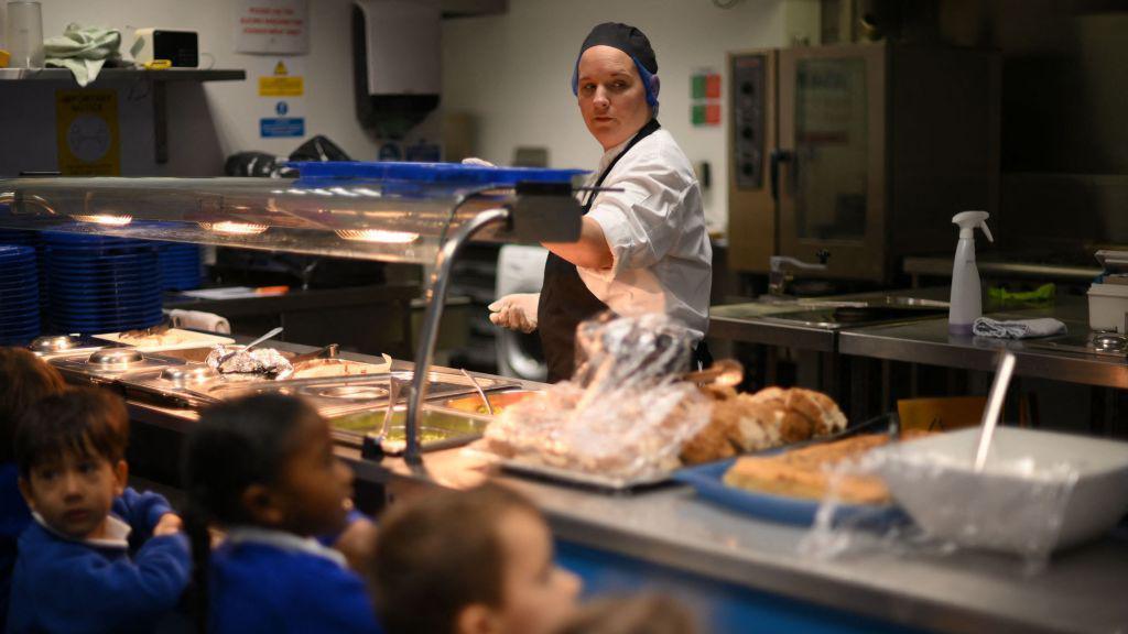 A canteen worker spoons food onto a blue tray. She wears gloves, a white top and dark apron and hat. The food is in trays under a hot lamp.