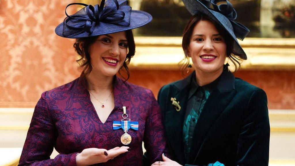 Georgia and Melissa Laurie dressed in smart maroon and green outfits at Bucking Palace. They are wearing dress hats while posing with a King's Gallantry medal on Georgia's chest.