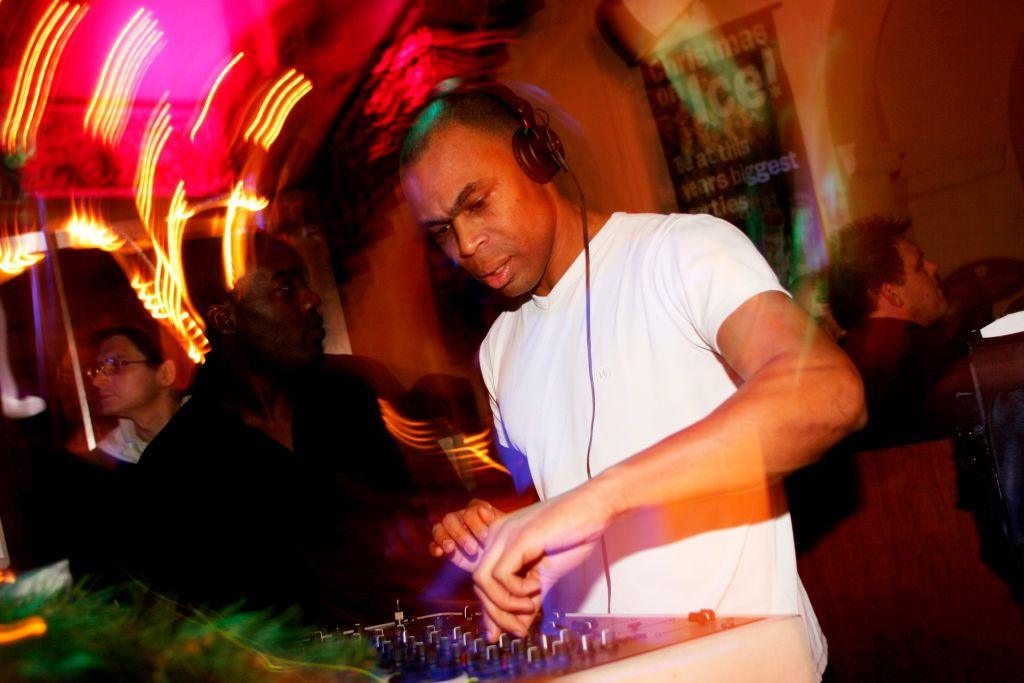 A photo blurred by flashing disco lights showing a man wearing headphones and a white T-shirt DJ-ing with people in the background at Batchwood Hall