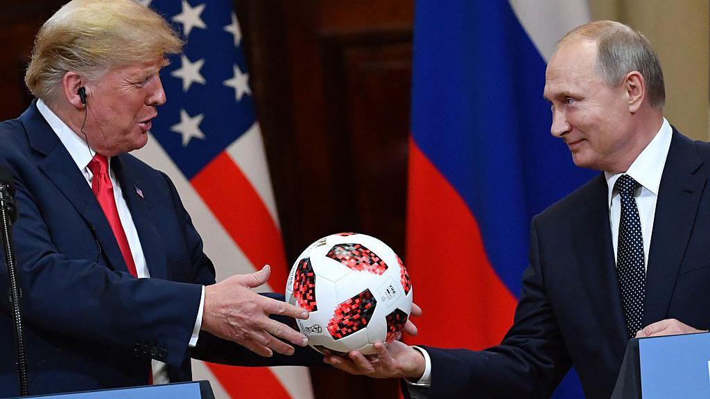 Vladimir Putin smiles as he hands a white and red football to Donald Trump during a meeting in Helsinki, Finland, in July 2018. Behind the pair are a US and a Russian flag. 