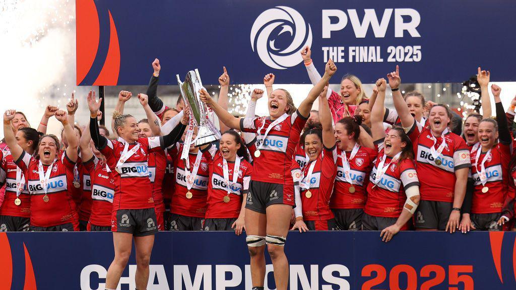 Gloucester-Hartpury players celebrate with the PWR trophy and a podium after winning the 2025 final