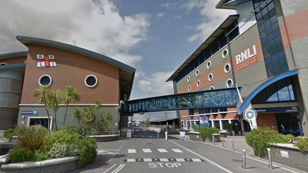 Two buildings connected by a glass bridge. One building has a sign up which reads: "RNLI College" and the other has the RNLI logo on it - a white and red flag.