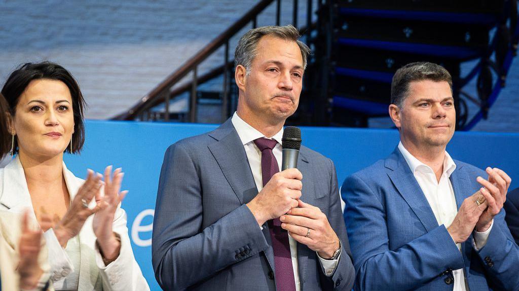 Alexander De Croo in the middle holding a microphone and looking sad