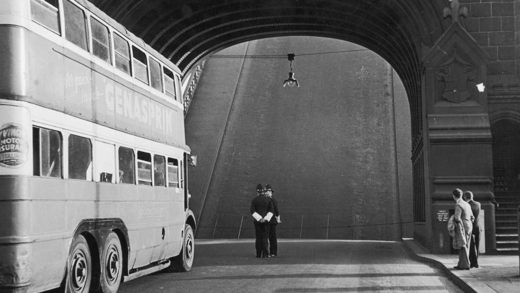 Archive black-and-white photo from 1946, showing a double-decker bus paused on Tower Bridge. The section of the road which raises to let boats through is up and looks like a steeply sloping wall in front of the bus. Two policemen chat in the road, wearing helmets and arm gaiters. Two men in suits, one carrying a coat, are on the pavement and are looking toward the section of raised bridge