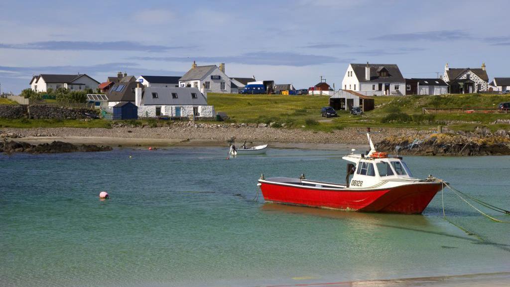 A small fishing boat with a red hull is tied up in a shallow expanse of turquoise water. Land in the background is dotted with white houses.