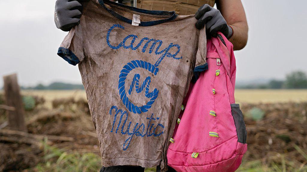 Someone holds up a very muddy t-shirt that has 'Camp Mystic' logo on it