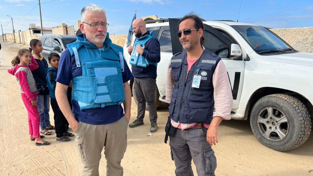 Picture of David Anderson in a blue flack jacket in Gaza he is standing with a fellow medic in front of a white UN 4x4.