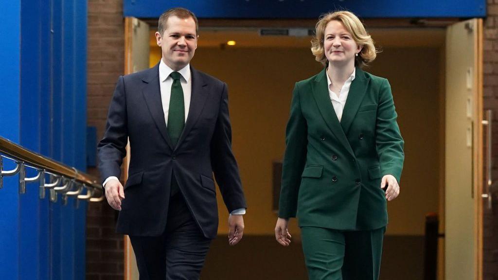 Katie Lam MP walks alongside Robert Jenrick. They are walking down a long corridor. Jenrick is wearing a dark suit, while Lam is wearing a brown suit.