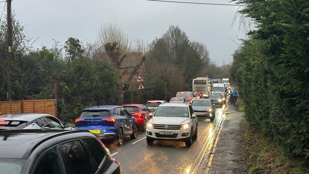 A traffic jam is pictured on a rural two lane road. A cyclist in the distance is using the pavement. 