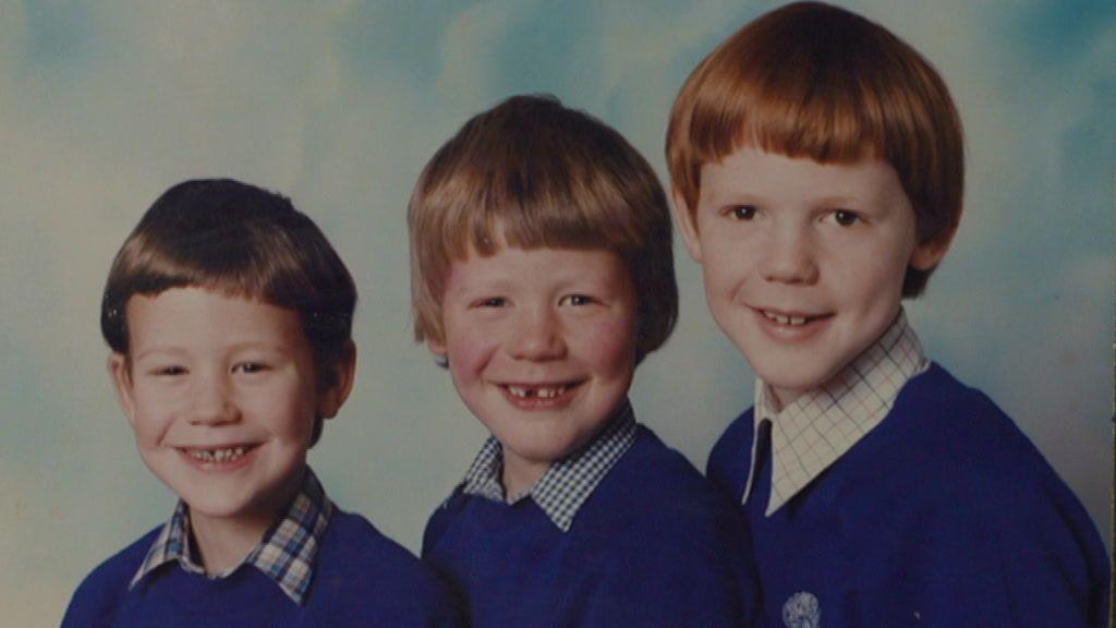 Three young boys, one with dark brown hair, one with light brown hair, and one with auburn, all wearing blue jumpers with different checked shirts poking out the collar.