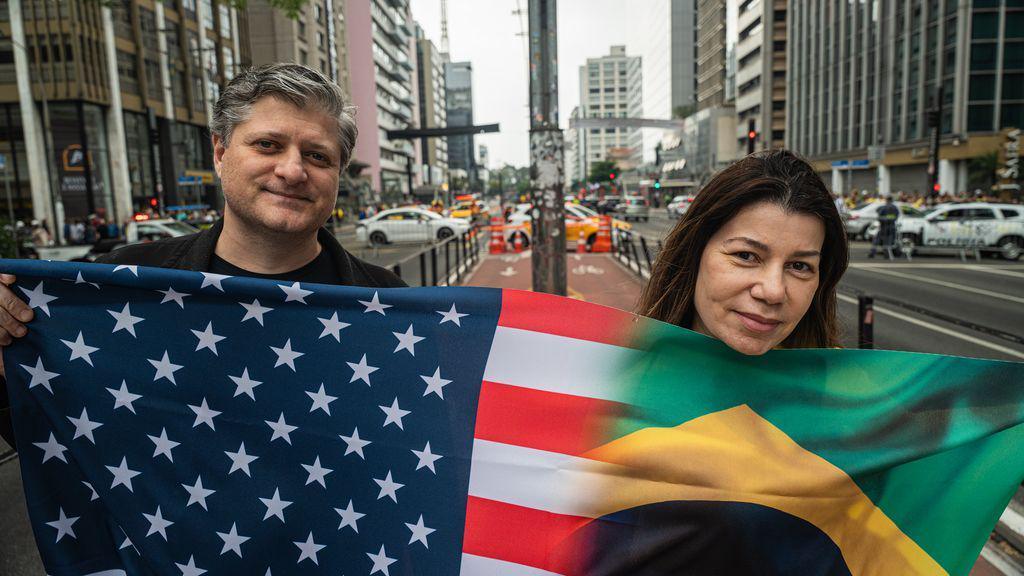 Bianca and a man hold up the flag, which bears the stars of the US flag on the left and the green and yellow of the Brazilian flag on the right. 