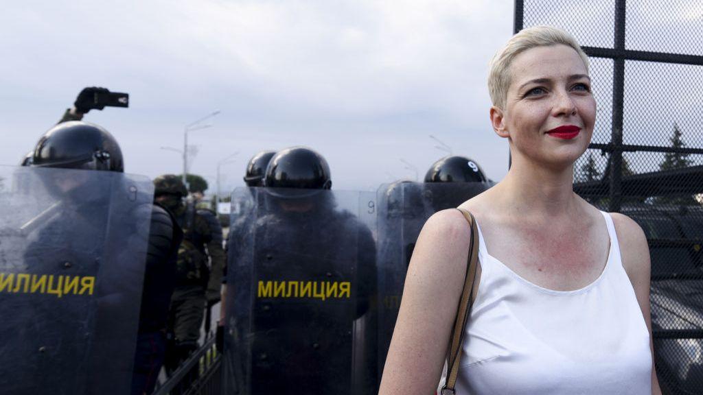 Maria, wearing a white singlet and with short blonde hair, smiles as she stands in front of officers in riot gear with sheilds.